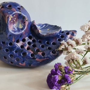 Puede incluir: Un pájaro de cerámica azul con agujeros y un esmalte rosa y amarillo. El pájaro está acostado de lado con un pequeño ramo de flores blancas a su lado.
