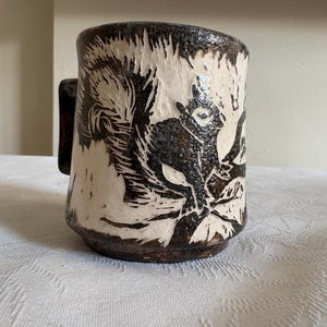 May include: A handcrafted ceramic mug with a dark brown exterior and a cream interior. The mug features a black and white illustration of a stylized animal. The handle is a dark brown colour.