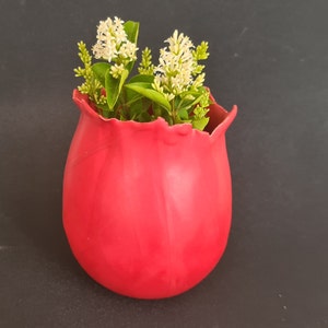 Può includere: Un vaso in ceramica rosso vivo con un bordo ondulato unico, pieno di fiori bianchi e foglie verdi.