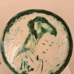 Puede incluir: Un plato redondo de cerámica con un esmalte verde y blanco. El plato presenta una imagen en relieve de la cara de una mujer de perfil. El cabello de la mujer está recogido hacia atrás y lleva un kimono japonés tradicional.