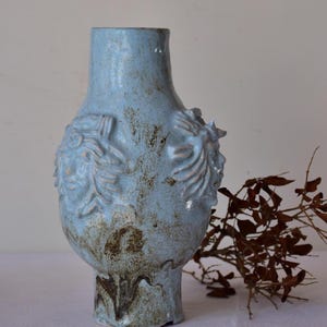 Könnte beinhalten: Hellblaue Keramikvase mit bauchigem Körper und schmalem Hals. Die Vase hat erhabene florale oder blattartige Designs und ein strukturiertes, gesprenkeltes Aussehen mit braunen Akzenten. Die Vase ist ein dekorativer Gegenstand.
