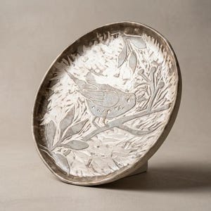 May include: A round ceramic plate with a textured, off-white surface featuring a dark brown, etched illustration of a bird perched on a branch surrounded by leaves. The plate has a raised, uneven rim and a dark brown finish along the outer edge.