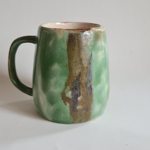 May include: A green ceramic mug with a brown and black glaze. The mug has a rounded shape and a handle.