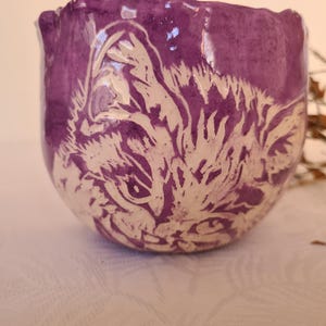 Puede incluir: Un cuenco de cerámica morado con un diseño de cara de gato blanco. La cara del gato está grabada en la superficie del cuenco. El cuenco está sobre una superficie blanca. Es un objeto decorativo.