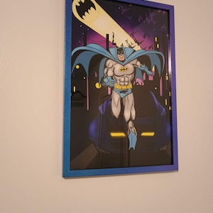 Puede incluir: Obra de arte enmarcada de Batman, con el superhéroe en una pose dinámica. La imagen muestra a Batman en azul y gris, sobre un fondo de paisaje urbano con un foco y la bat-señal. El marco es azul.