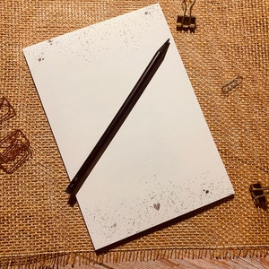 May include: A blank white notepad with a black pencil on top of it. The notepad has a subtle speckled pattern and two small hearts in the bottom right corner. The notepad is on a brown burlap surface.