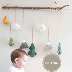 May include: A handmade baby mobile with a wooden branch, orange string, and crocheted ornaments. The ornaments include a crescent moon, clouds, trees, and mountains. The mobile is hanging from the ceiling and a baby is looking at it.