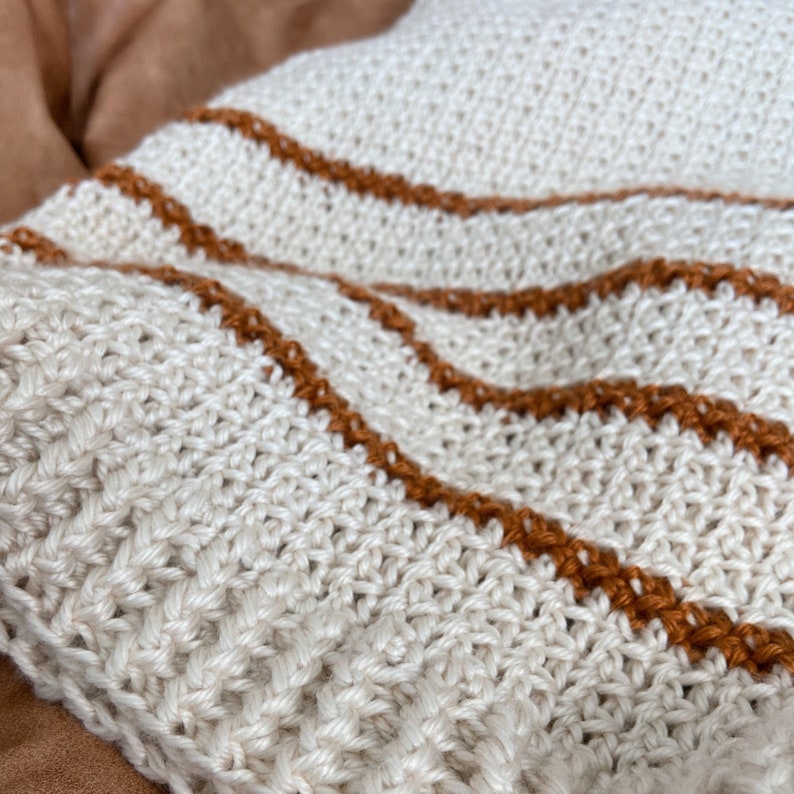 May include: A white crocheted throw blanket with brown stripes. The blanket is made with a textured stitch and has a soft, cozy feel.