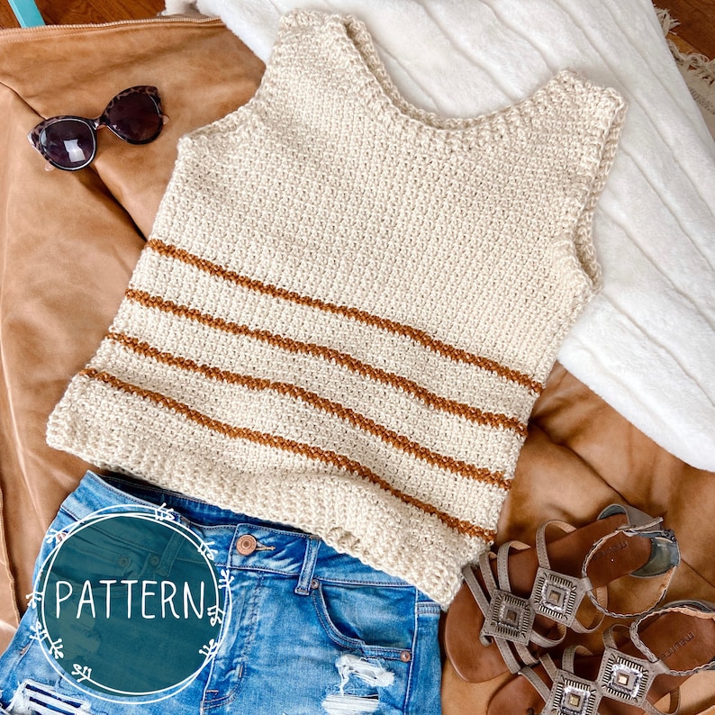 May include: A crocheted sleeveless top with brown and white stripes. The top is laying on a brown leather surface with a pair of jeans and sandals.