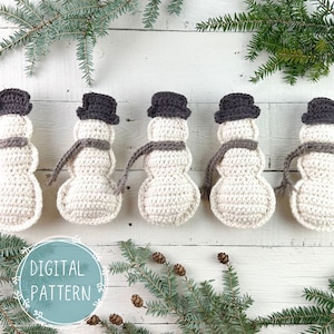 May include: Five crocheted snowmen ornaments with brown hats and scarves, arranged in a row on a white wooden background. The snowmen are made with white yarn and have a simple, classic design. The background features a sprig of evergreen branches with pine cones.