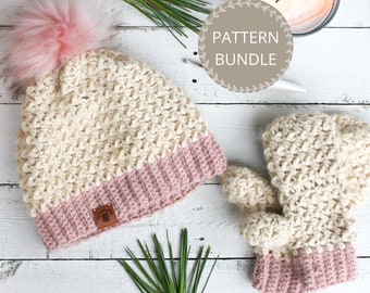 Könnte beinhalten: Ein Häkelset aus Mütze und passenden Fäustlingen in Creme und Rosa mit einem flauschigen Bommel auf der Mütze. Das Set liegt auf einer weißen Holzoberfläche mit Kiefernzweigen.