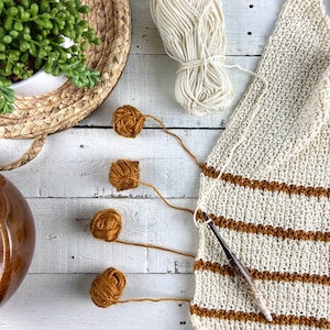 May include: A close-up of a crocheted blanket with brown and white stripes. Four balls of yarn in various shades of brown are on a white surface with a crochet hook.