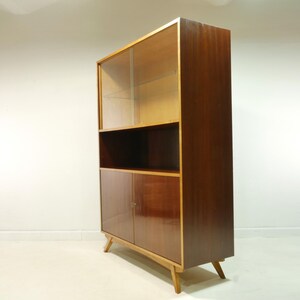 May include: A mid-century modern display cabinet with a dark wood finish. The upper section has a glass door, the middle has an open shelf, and the bottom has two doors. The cabinet is supported by angled wooden legs.