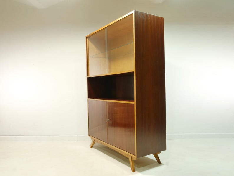May include: A mid-century modern display cabinet with a dark wood finish. The upper section has a glass door, the middle has an open shelf, and the bottom has two doors. The cabinet is supported by angled wooden legs.