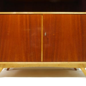 May include: A mid-century modern wooden cabinet with a light-colored frame and legs. The cabinet doors are a rich, reddish-brown color, with a small silver keyhole. The cabinet has a top shelf.