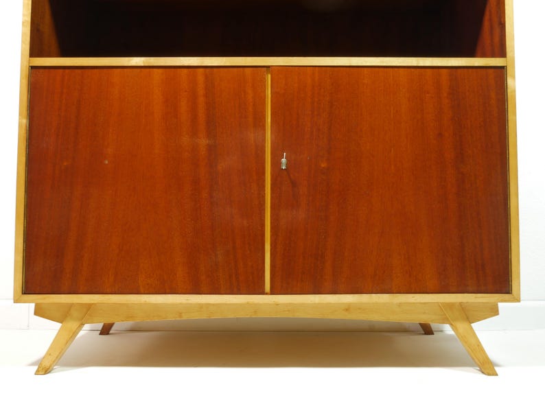 May include: A mid-century modern wooden cabinet with a light-colored frame and legs. The cabinet doors are a rich, reddish-brown color, with a small silver keyhole. The cabinet has a top shelf.