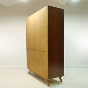 May include: A mid-century modern wooden cabinet with a light wood front and a dark wood side. The cabinet has a rectangular shape and stands on four angled legs. The cabinet is likely used for storage.