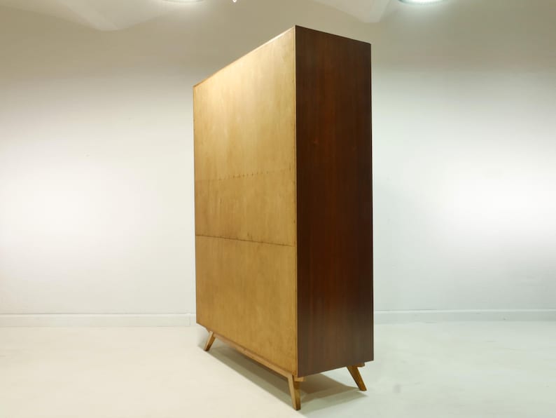 May include: A mid-century modern wooden cabinet with a light wood front and a dark wood side. The cabinet has a rectangular shape and stands on four angled legs. The cabinet is likely used for storage.
