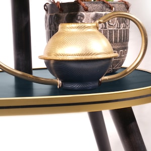 Op de afbeelding: Een gouden en blauwe gieter met een koperen tuit en handvat staat op een donkerblauwe ovale tafel met een gouden rand. De tafel heeft drie zwarte poten.