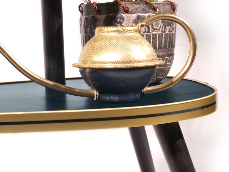 Op de afbeelding: Een gouden en blauwe gieter met een koperen tuit en handvat staat op een donkerblauwe ovale tafel met een gouden rand. De tafel heeft drie zwarte poten.