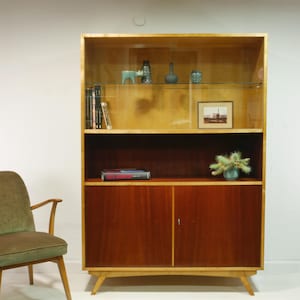 May include: A mid-century modern wooden bookcase with a light wood frame and dark wood doors. The bookcase has glass shelves displaying decorative items and books. A green upholstered chair sits to the left.