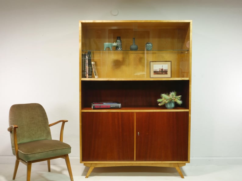 May include: A mid-century modern wooden bookcase with a light wood frame and dark wood doors. The bookcase has glass shelves displaying decorative items and books. A green upholstered chair sits to the left.