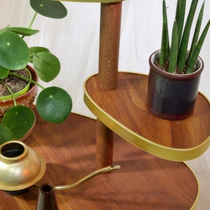 May include: A three-tiered plant stand made of wood with a gold trim. The stand has two oval-shaped shelves and a round base. There are two potted plants on the stand, one with green leaves and the other with long, thin green leaves. A gold watering can is on the bottom shelf.