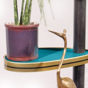 Op de afbeelding: Een tealblauw rek met een gouden rand en zwarte afwerking zit op een zwarte metalen standaard. Een plant in een pot met groene bladeren staat op het rek. Een gouden metalen vogelsculptuur staat op het rek.