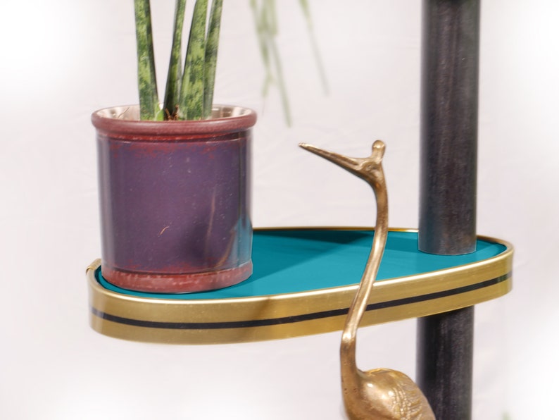 Op de afbeelding: Een tealblauw rek met een gouden rand en zwarte afwerking zit op een zwarte metalen standaard. Een plant in een pot met groene bladeren staat op het rek. Een gouden metalen vogelsculptuur staat op het rek.
