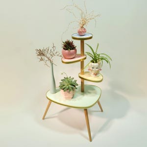 Op de afbeelding: Een witte plantenstandaard met vier niveaus, elk met een lichtblauwe of roze rand. De standaard heeft een houten basis en poten. De bovenste laag heeft een kleine roze vaas met gedroogde bloemen. De tweede laag heeft een roze pot met een groene succulent. De derde laag heeft een witte pot met een groene plant. De onderste laag heeft een roze pot met een groene succulent.
