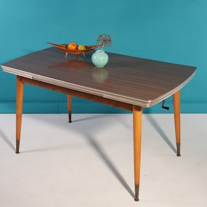 Può includere: Un tavolo in laminato marrone e bianco con un bordo argentato e una manovella sul lato. Il tavolo ha quattro gambe in legno con punte in ottone. Un piccolo vaso blu con un'arrangiamento floreale si trova sul tavolo.