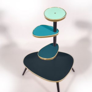 Op de afbeelding: Een drietraps plantenstandaard met een zwart metalen frame en teal- en lichtblauwe glazen planken. De standaard heeft een unieke, organische vorm en is perfect om planten of andere decoratieve items te presenteren.