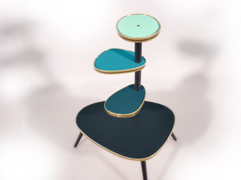 Op de afbeelding: Een drietraps plantenstandaard met een zwart metalen frame en teal- en lichtblauwe glazen planken. De standaard heeft een unieke, organische vorm en is perfect om planten of andere decoratieve items te presenteren.