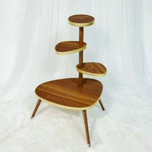 May include: A mid-century modern plant stand with four tiers. The stand is made of wood with a dark brown finish and gold accents. The top tier is round, and the other three tiers are oval shaped.