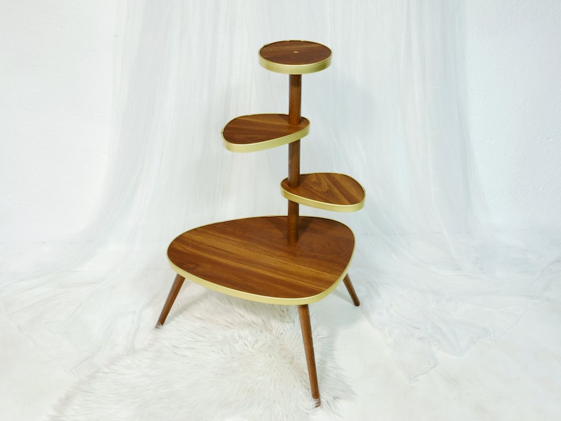 May include: A mid-century modern plant stand with four tiers. The stand is made of wood with a dark brown finish and gold accents. The top tier is round, and the other three tiers are oval shaped.