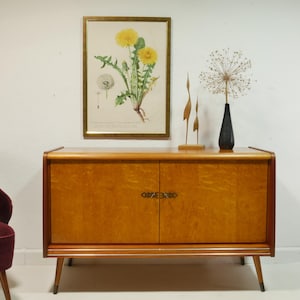 Può includere: Un mobile in legno di stile moderno di metà secolo con due ante e una finitura in legno chiaro. Il mobile ha ferramenta in ottone e poggia su quattro gambe. Una stampa botanica incorniciata di dente di leone e una scultura metallica decorativa sono sopra il mobile.