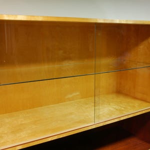 May include: A light-colored wooden display cabinet with glass sliding doors. The cabinet has two shelves and a lower open shelf. The wood has a natural finish, and the glass doors are clear, offering visibility of the interior.