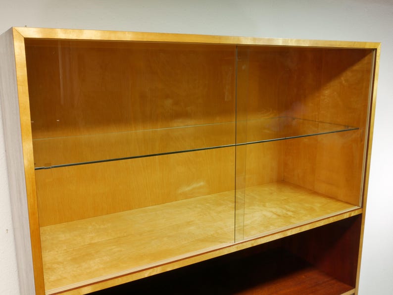 May include: A light-colored wooden display cabinet with glass sliding doors. The cabinet has two shelves and a lower open shelf. The wood has a natural finish, and the glass doors are clear, offering visibility of the interior.