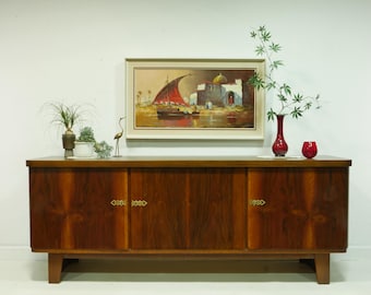 Può includere: Credenza in legno di metà secolo con quattro ante e ferramenta in ottone. La credenza è sormontata da un dipinto incorniciato di una barca e di architetture mediorientali. Piante decorative e un vaso rosso sono in cima. Misura circa 150 cm di lunghezza.