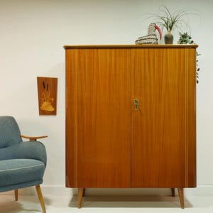 Könnte beinhalten: Ein Vintage-Holzschrank in warmem, rötlich-braunem Farbton. Der Schrank hat zwei Türen mit einem goldenen Griff und steht auf vier konischen Beinen. Ein blauer Sessel und dekorative Gegenstände sind ebenfalls zu sehen.