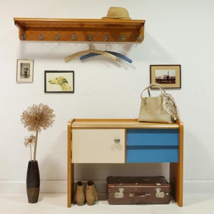 Könnte beinhalten: Ein Vintage-Eingangsset mit einer Garderobe aus Holz, einem kleinen Schrank mit blauen und cremefarbenen Schubladen und einem Koffer. Das Set umfasst gerahmte Bilder, einen Hut, eine Handtasche und eine Vase mit getrockneten Blumen. Das Farbschema ist neutral mit Blautönen.