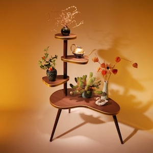 May include: A mid-century modern plant stand with multiple levels, holding various plants and decorative items. The stand is made of dark wood with gold trim and has a unique, curved shape. It features a variety of plants and vases.