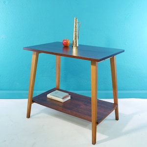 Op de afbeelding: Een donkerhouten bijzettafel met een onderplank. De tafel heeft een licht houten frame en een enkele rode appel op de bovenste plank.