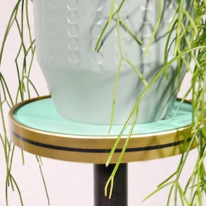Op de afbeelding: Een turquoise plantenstandaard met een gouden rand en een zwarte basis. Een turquoise pot met een getextureerd oppervlak staat op de standaard.