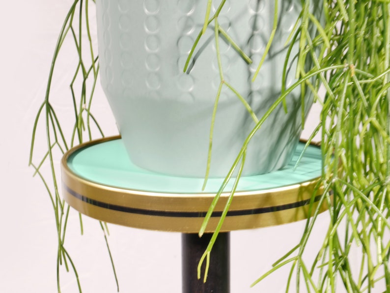 Op de afbeelding: Een turquoise plantenstandaard met een gouden rand en een zwarte basis. Een turquoise pot met een getextureerd oppervlak staat op de standaard.