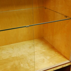 May include: A light-colored wooden display cabinet with glass shelves and a glass door. The cabinet has a simple, minimalist design with clean lines and a natural wood grain finish. The shelves are supported by metal brackets.