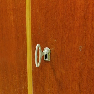 May include: Close-up of a wooden cabinet door with a silver key inserted into the keyhole. The door is a warm brown color with a vertical yellow stripe. The key is in the center of the frame.