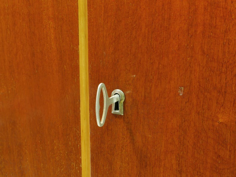 May include: Close-up of a wooden cabinet door with a silver key inserted into the keyhole. The door is a warm brown color with a vertical yellow stripe. The key is in the center of the frame.