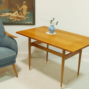 Op de afbeelding: Een houten bijzettafel in mid-century stijl met een rechthoekig blad en taps toelopende poten. De tafel heeft een lichtbruine afwerking en messingkleurige uiteinden op de poten. Een blauwe fauteuil en een schilderij staan op de achtergrond.