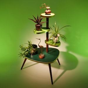 Op de afbeelding: Een groene en gouden plantenstandaard met vier niveaus. De standaard heeft een unieke, gebogen vorm en beschikt over een verscheidenheid aan planten en decoratieve items, waaronder een klein vogelsculptuur.